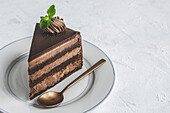 Piece of yummy sweet chocolate cake decorated with cream served on ceramic plate with golden spoon on white tablecloth