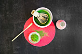 Top view of healthy matcha tea and dried powder with wooden chasen and chashaku on bright round stand on black table at sunlight