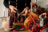 Crop unrecognizable female florist standing and cutting twigs of fir while arranging Christmas bouquet on wooden table