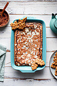 Top view of tasty baked brownie with almond flakes in baking tray with cookies placed on wooden table with cocoa powder