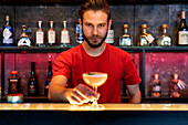 Cheerful barman serving glass with alcohol cocktail on counter in bar and looking at camera