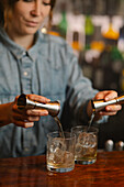 Crop unrecognizable barkeeper pouring alcohol drinks from jiggers into glasses with ice cubes while preparing cocktails at car counter on blurred background