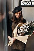 Positive woman in stylish clothes and beret standing with creative bouquet with twigs of cotton and fir in light room