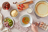 Top view of unrecognizable woman using whisk to beat raw chicken eggs in bowl near fresh ingredients and crust while preparing quiche in kitchen at home