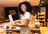 Young smiling African American woman in uniform and glasses with curly hair looking at camera checking goods in glass boxes and using laptop while working in zero waste grocery store