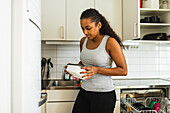 Calm ethnic lady in light kitchen standing near dishwasher and opened white kitchen cabinet filled with kitchenware while closing food plastic container in daylight