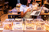 Cropped unrecognizable young African American woman in uniform and glasses with curly hair checking goods in glass boxes and using laptop while working in zero waste grocery store