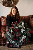 Fröhliche Frau in Freizeitkleidung sitzt mit dekorativem Weihnachtskranz in den Händen auf dem Sofa und schaut in die Kamera