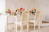 Chairs placed neat table with white tablecloth and flowers against window in sunlit room with shabby walls during wedding celebration