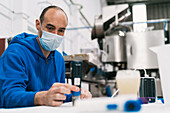 Male quality control specialist in sterile mask studying alcoholic liquid at table against professional equipment in factory