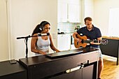 Ruhige hispanische Frau mit Kopfhörern, die E-Piano spielt und in ein Mikrofon singt, während sie mit einem Freund, der Akustikgitarre spielt, tagsüber zu Hause Musik macht