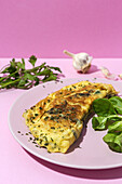 Tasty omelette on plate against fresh parsley sprigs with garlic cloves on pink background