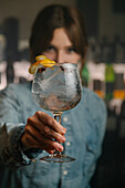 Soft focus female bartender looking at camera while serving wineglass with gin and tonic cocktail decorated with orange zest on wooden counter in bar