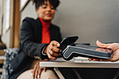 Crop African American female client putting cellphone on terminal in hand of waitress and paying for order in cafe