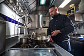 Plump bearded man in dark uniform putting frying pan on cooktop while cooking dish in contemporary restaurant kitchen