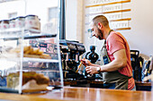 Side view of focused male barista in apron cleaning coffee machine with brush while standing near counter with desserts in coffee house