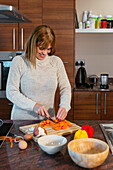 Smiling adult female in sweater cutting carrots wooden cutting board at counter with built in stovetop and bowls while making dinner