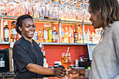 Cheerful black female barkeeper serving glass of refreshing cocktail for client in bar