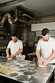 Mature bearded ethnic bakers forming bread from dough at table with flour and bowl in bakery