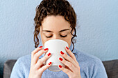 Calm female with curly hair drinking hot beverage from white cup while sitting on couch at wall in light room