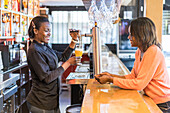Seitenansicht einer schwarzen Barkeeperin, die einer am Tresen sitzenden Frau Bier in ein Glas gießt