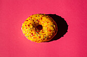 Draufsicht auf einen mit gelbem Zucker bestrichenen Donut mit bunten Kugeln auf rosa Hintergrund