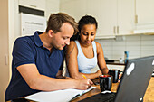 Smiling young multiracial couple using gadgets and devices while sitting at counter in kitchen and studying