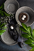 From above place setting for Easter dinner with ceramic plates easter eggs in nest surrounded by Italian ruscus and cushion baby's-breath leaves