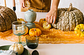 Crop unrecognizable female designer in apron creating composition with various pumpkins and glassware for Halloween party