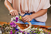 Crop anonymous female florist creating wreath from fresh colorful flowers while working at table in shop
