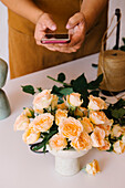 From above of crop unrecognizable floral designer in apron using smartphone while taking photo of delicate roses composition arranged on table in light studio