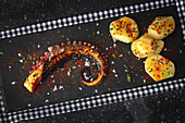 Top view of fried octopus tentacle and pieces of potato served with spices on black board on table