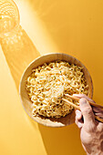 Top view of crop anonymous person with wooden chopsticks eating fresh noodles in bowl placed on yellow background with glass