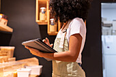 Cropped side view of unrecognizable black female vendor in uniform with tablet smiling and inspecting pasta in glass container during work in zero waste grocery store looking away