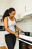 African American housewife wearing black trousers and gray top cooking meal in saucepan on stove in light kitchen