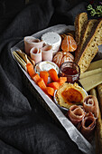 From above brunch box with assorted sliced meats various types of cheese and crispbreads arranged near ripe cup kiwi sweet strawberries and peeled mandarin near jam in glass jar on wooden table near napkin