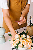 From above of crop unrecognizable floral designer in apron using smartphone while taking photo of delicate roses composition arranged on table in light studio