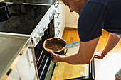 From above side view of adult male cooker in light kitchen opening oven door and putting baking form filled with chocolate cake batter inside