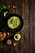 Top view of bowl of fresh guacamole placed near avocado and chips on weathered lumber tabletop