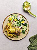 Top view of plate with omelette roll and radish and cucumber salad with herbs placed near artichoke and napkin on gray table