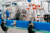 Side view of male coworkers in sterile masks speaking against mash boiler and lauter tun while looking at each other during pandemic