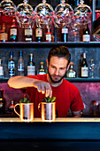 Content male bartender garnishing Moscow mule cocktails with mint leaves served in copper cups on counter in bar