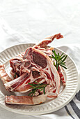 From above of uncooked lamb chops served on white ceramic plate with fresh green rosemary on table in light kitchen