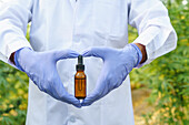 Crop anonymous female scientist wearing white robe and latex gloves demonstrating glass bottle with liquid drug