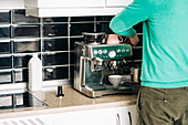 Back view of crop unrecognizable male preparing coffee with modern espresso machine at table in house kitchen