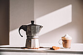 Metal geyser coffee maker placed on table with aromatic foamy latte macchiato served in Irish glass with heart shape cookie in kitchen against white wall on sunny day