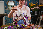 Focused female florist making flower wreath with ribbon bow while working in floral shop