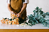 Anonymous female floral designer in apron standing at table and creating floral composition with blooming roses in workshop