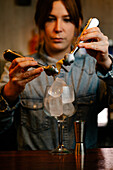 Soft focus of crop female barkeeper putting with tongs lemon zest into glass with ice cubes while making cocktail in bar