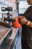 Abgeschnittene unerkennbare afroamerikanische Barkeeperin, die Milch in einem Metallkrug aufschlägt, während sie ein köstliches Getränk in einer Kaffeemaschine zubereitet